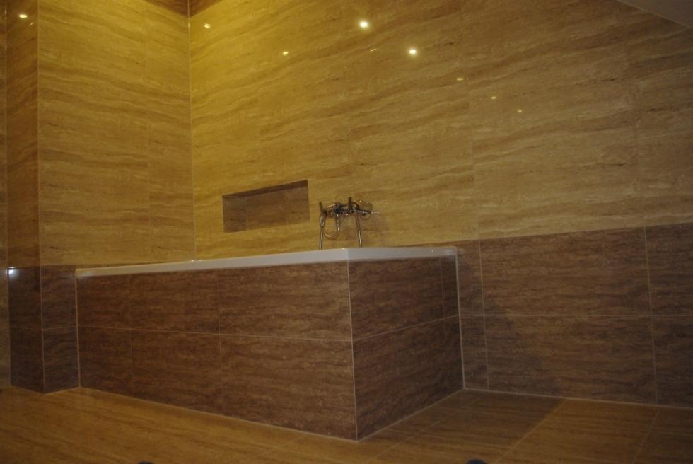 Wykończona łazienka z wanną obłożoną ciemnobrązowymi płytkami imitującymi drewno, ściany wyłożone beżowymi płytkami z efektem marmuru, w ścianie wnęka, bateria wannowa chromowana.
