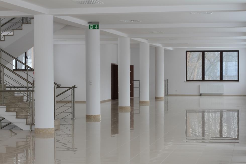 Jasne, przestronne wnętrze z polerowaną podłogą, białymi kolumnami, schodami z metalową balustradą i oknem z widokiem na drzewa.