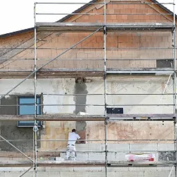 Dwukondygnacyjny budynek w trakcie prac elewacyjnych, otoczony rusztowaniem, z widocznymi fragmentami ocieplenia i pracującym na rusztowaniu mężczyzną w białym kombinezonie.