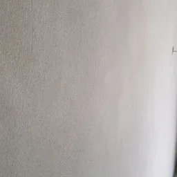 Świeżo nałożony, jasnoszary tynk na ścianie wewnętrznej, widoczna delikatna faktura i niewielki fragment podłogi. Mały ślad po gwoździu na ścianie.