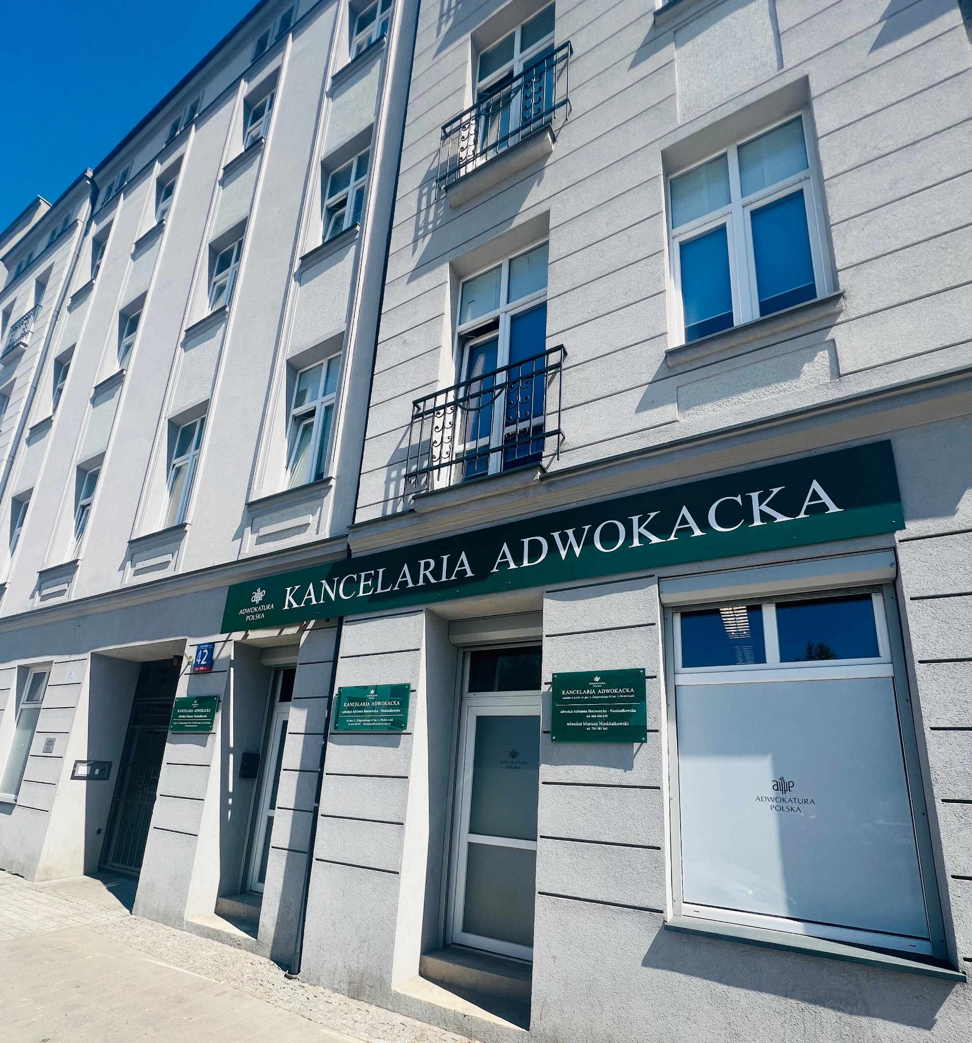 Fasada kamienicy z szyldem 'Kancelaria Adwokacka' w zielono-białej kolorystyce, tabliczki informacyjne po obu stronach wejścia, numer budynku 42, błękitne niebo w tle.