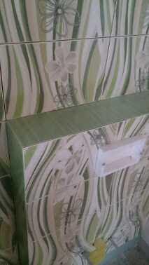 Ściana wyłożona płytkami ceramicznymi w odcieniach zieleni i bieli z motywem kwiatowym, widoczna półka z tego samego materiału i fragment białej umywalki.