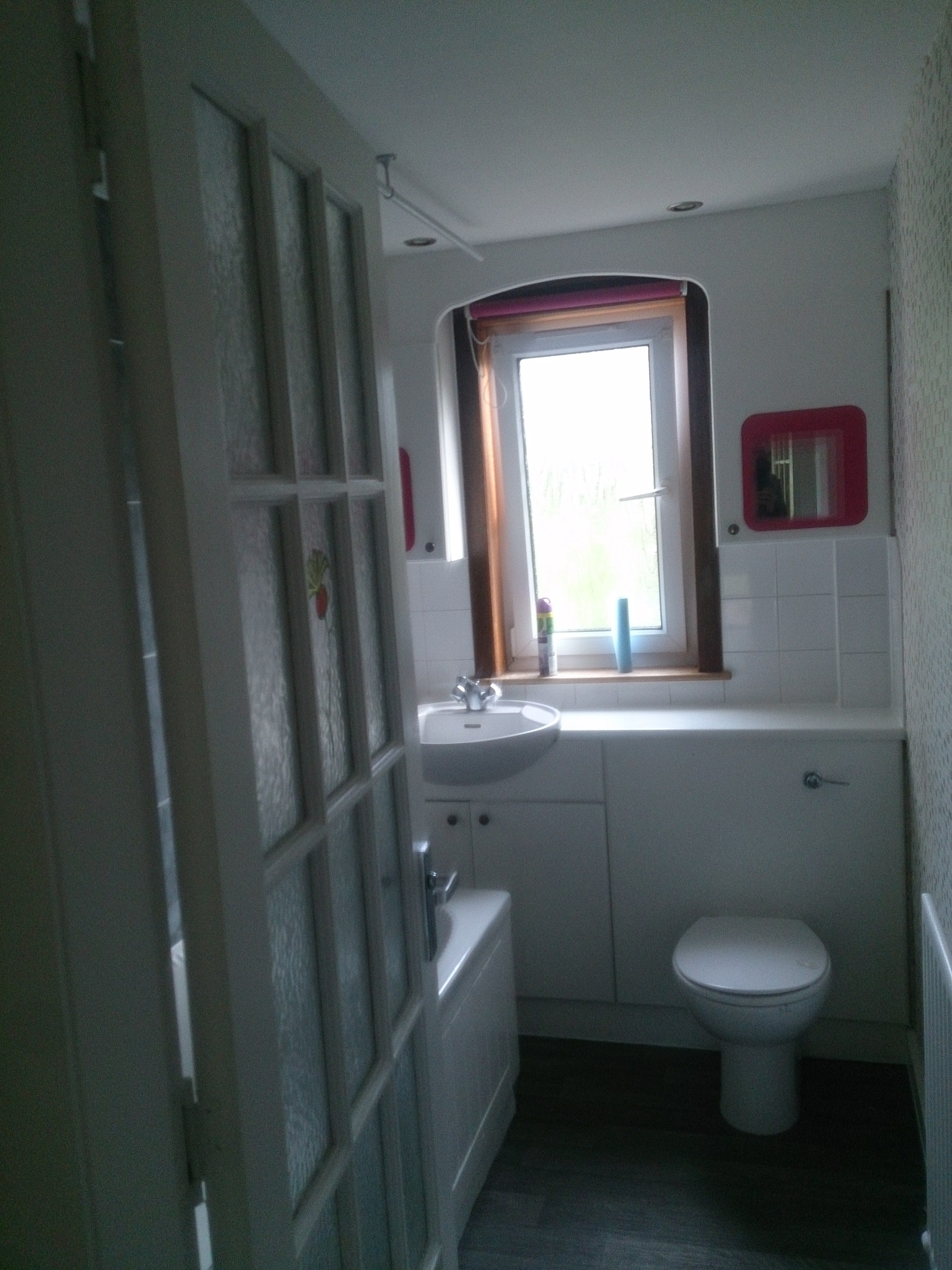 Widok na małą łazienkę z umywalką w zabudowie, toaletą i grzejnikiem, widzianą przez częściowo otwarte, przeszklone drzwi.