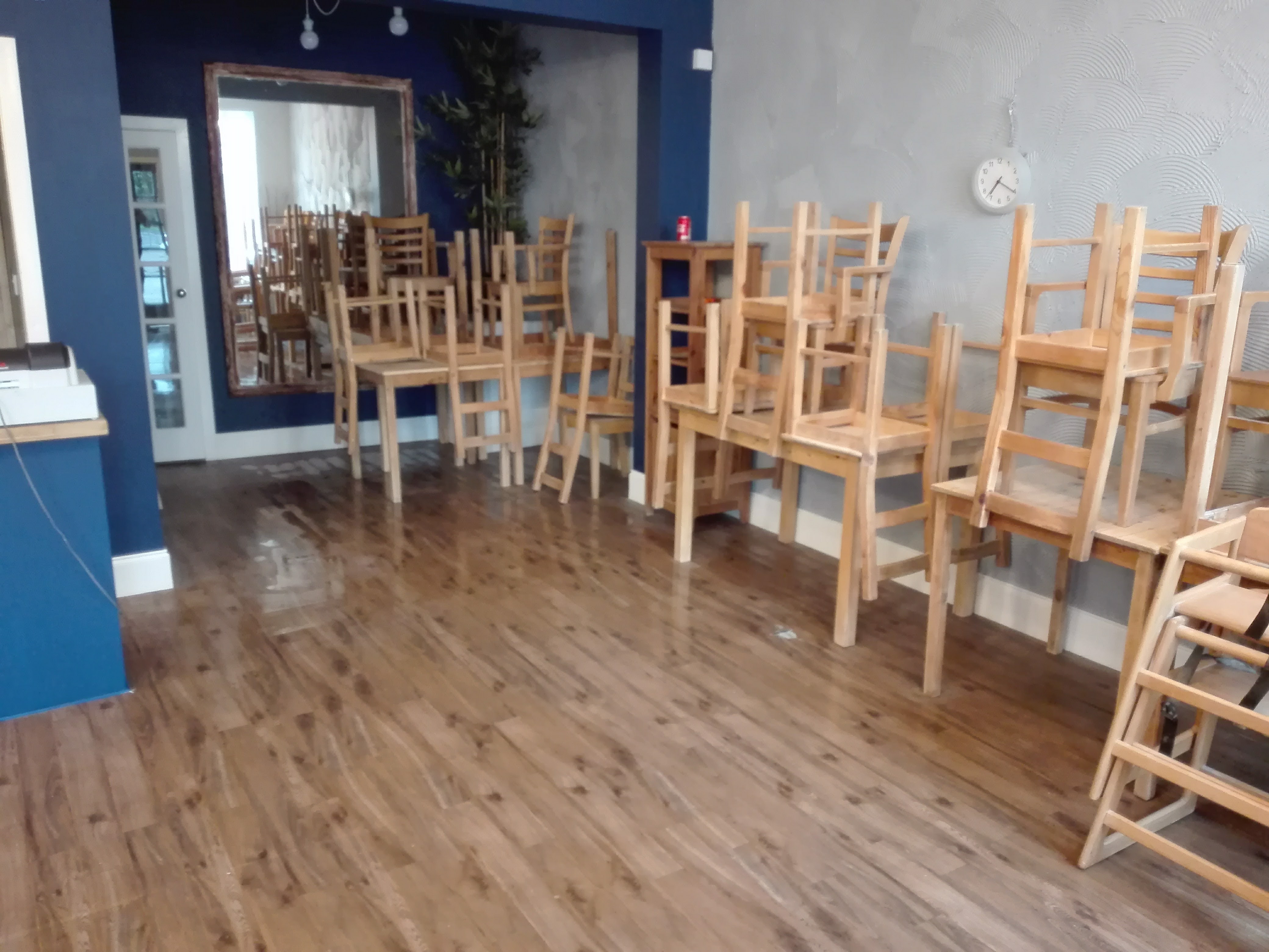 Pusta sala z parkietem, krzesła i stoły ustawione w stosy, przygotowane do malowania ścian w odcieniach granatu i szarości, widoczny zegar na ścianie.