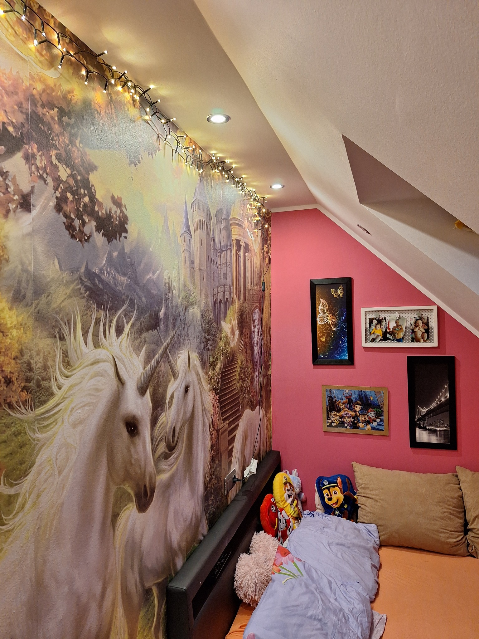 Pokój na poddaszu z tapetą przedstawiającą jednorożce i zamek, oświetlony girlandą lampek, z różową ścianą ozdobioną ramkami i pluszakami na łóżku.