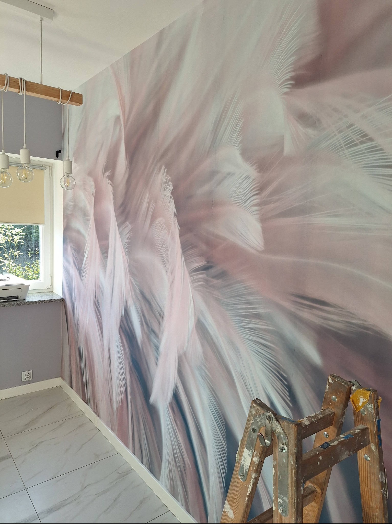 Pokój po tapetowaniu z motywem piór, widoczna drabina malarska i nowoczesna lampa wisząca.
