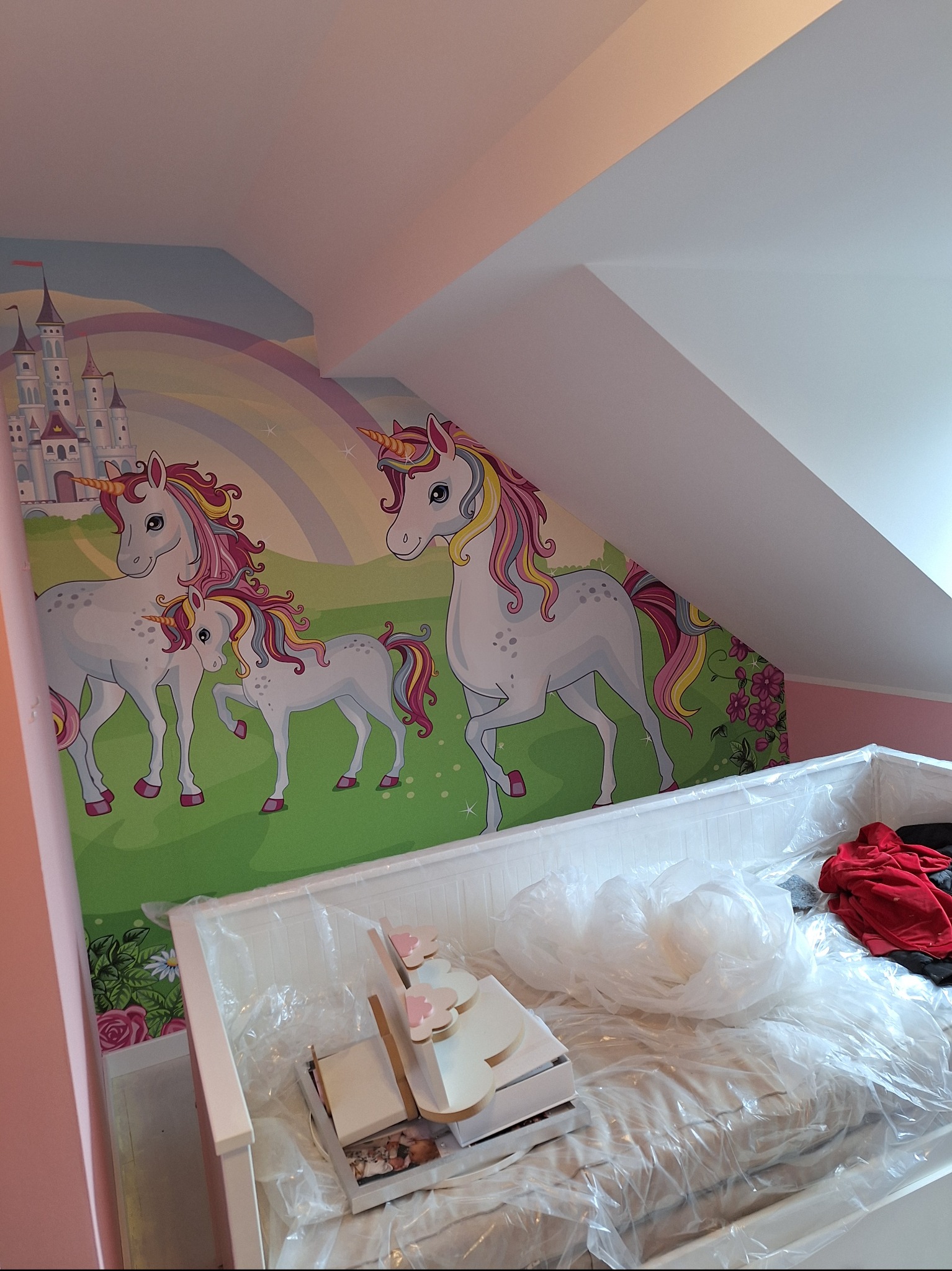Pokój dziecięcy z tapetą w jednorożce i tęczę, łóżko zabezpieczone folią malarską, widoczne akcesoria do pokoju.