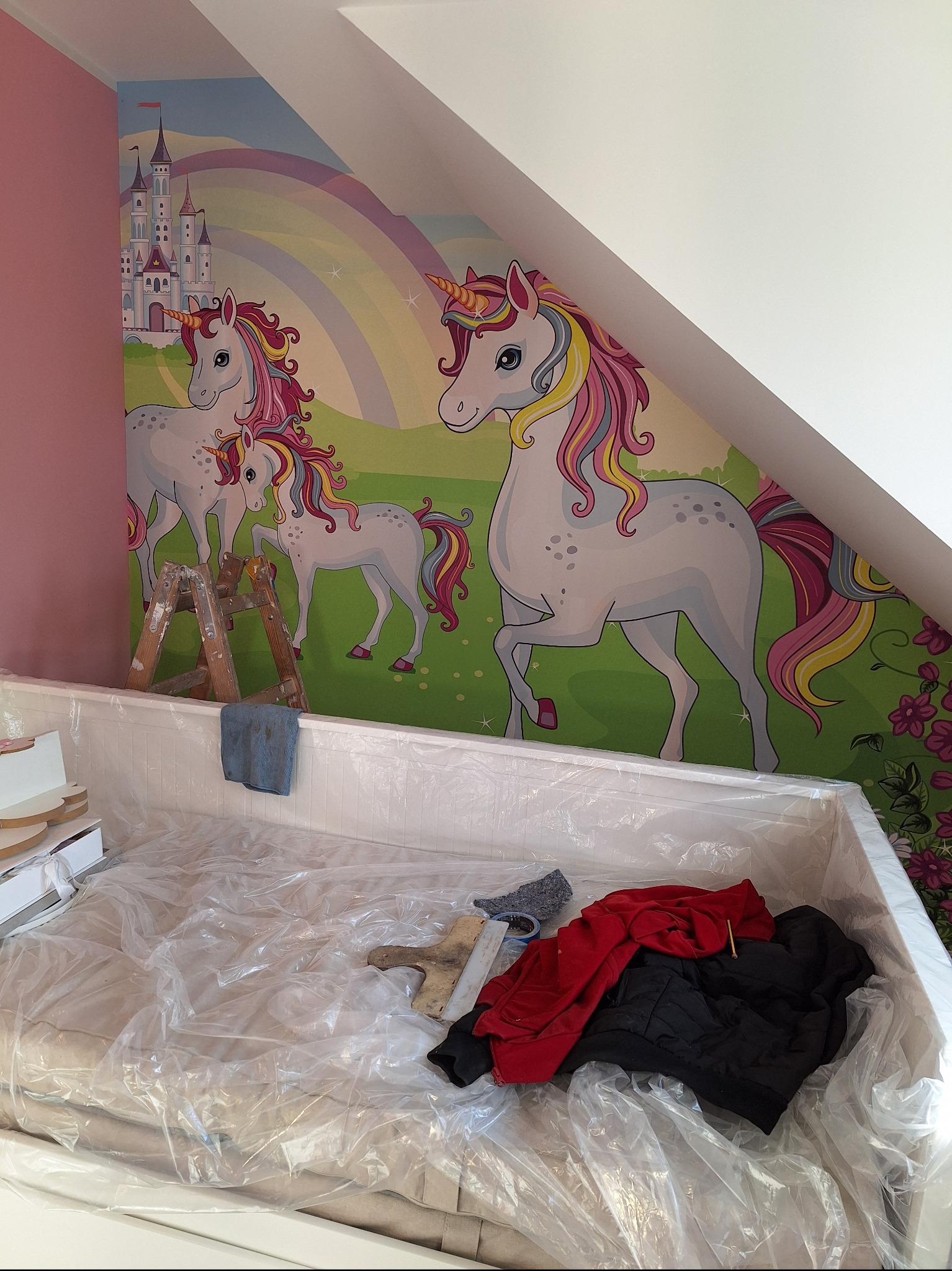 Pokój dziecięcy w trakcie dekorowania: na ścianie fototapeta z jednorożcami i zamkiem, drabina malarska, zabezpieczone meble folią, ubrania i narzędzia malarskie w kuwecie.