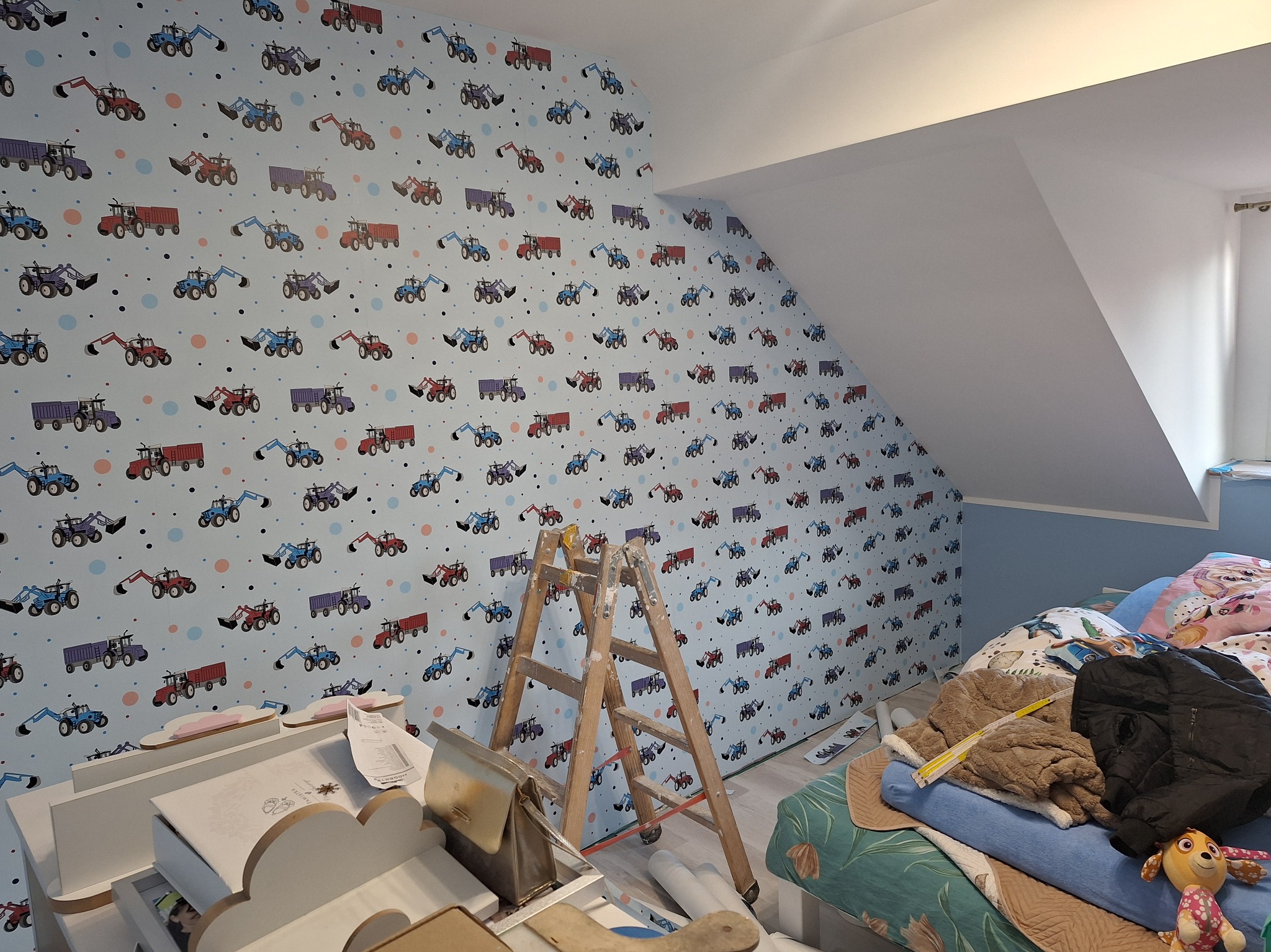Pokój dziecięcy w trakcie tapetowania: ściana pokryta tapetą w traktory, drabina malarska, zwinięte rolki tapety, zabawki na łóżku w tle.