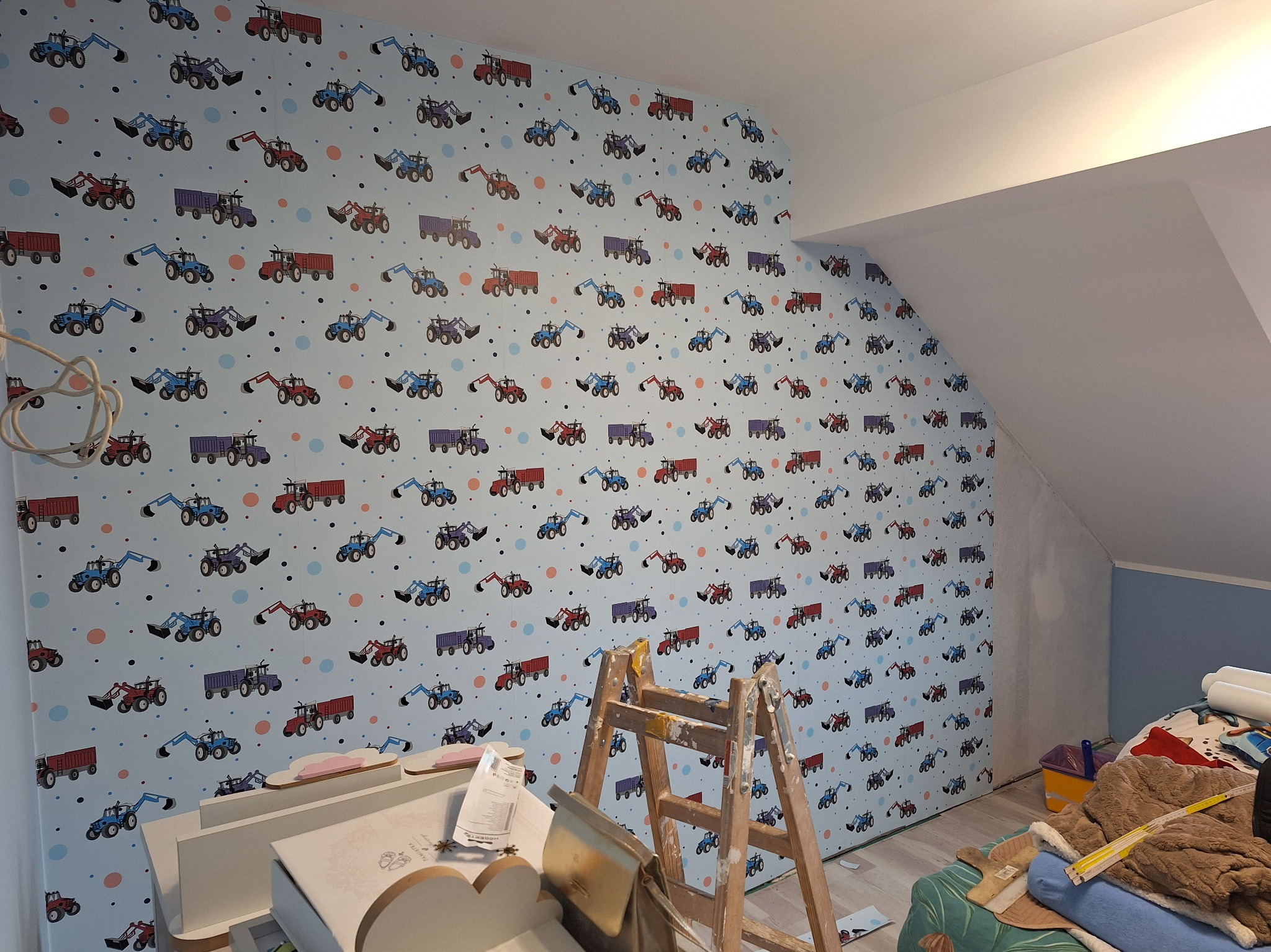 Pokój dziecięcy w trakcie wykańczania, ściana pokryta tapetą w traktory, drabina malarska oparta o ścianę, widoczne materiały wykończeniowe.