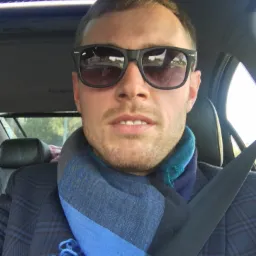 Mężczyzna w okularach przeciwsłonecznych i niebieskim szaliku siedzi w samochodzie z otwartym szyberdachem, zapięty pasem bezpieczeństwa.