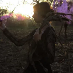 Kobieta w koronkowej sukience i z gałęziami na ramieniu trzyma w dłoni kulkę z fioletowym dymem na tle pola i zachodzącego słońca.