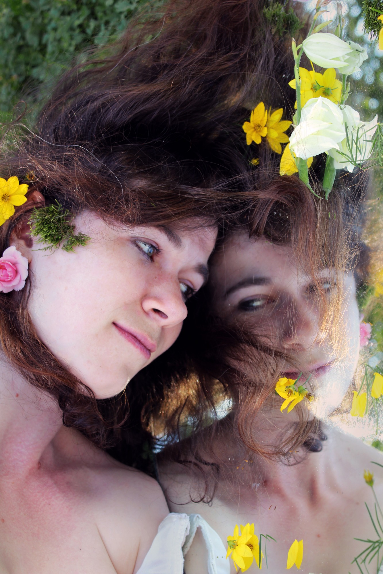 Portret kobiety z brązowymi włosami, ozdobionymi kwiatami i mchem, leżącej na trawie w Bochni, z odbiciem w lustrze. Romantyczne ujęcie z bliska.