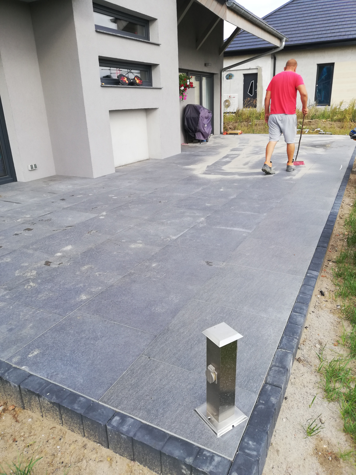 Szara kostka brukowa na tarasie domu jednorodzinnego, czyszczona przez mężczyznę z użyciem miotły, widoczny słupek oświetleniowy, obrzeża z kostki brukowej.