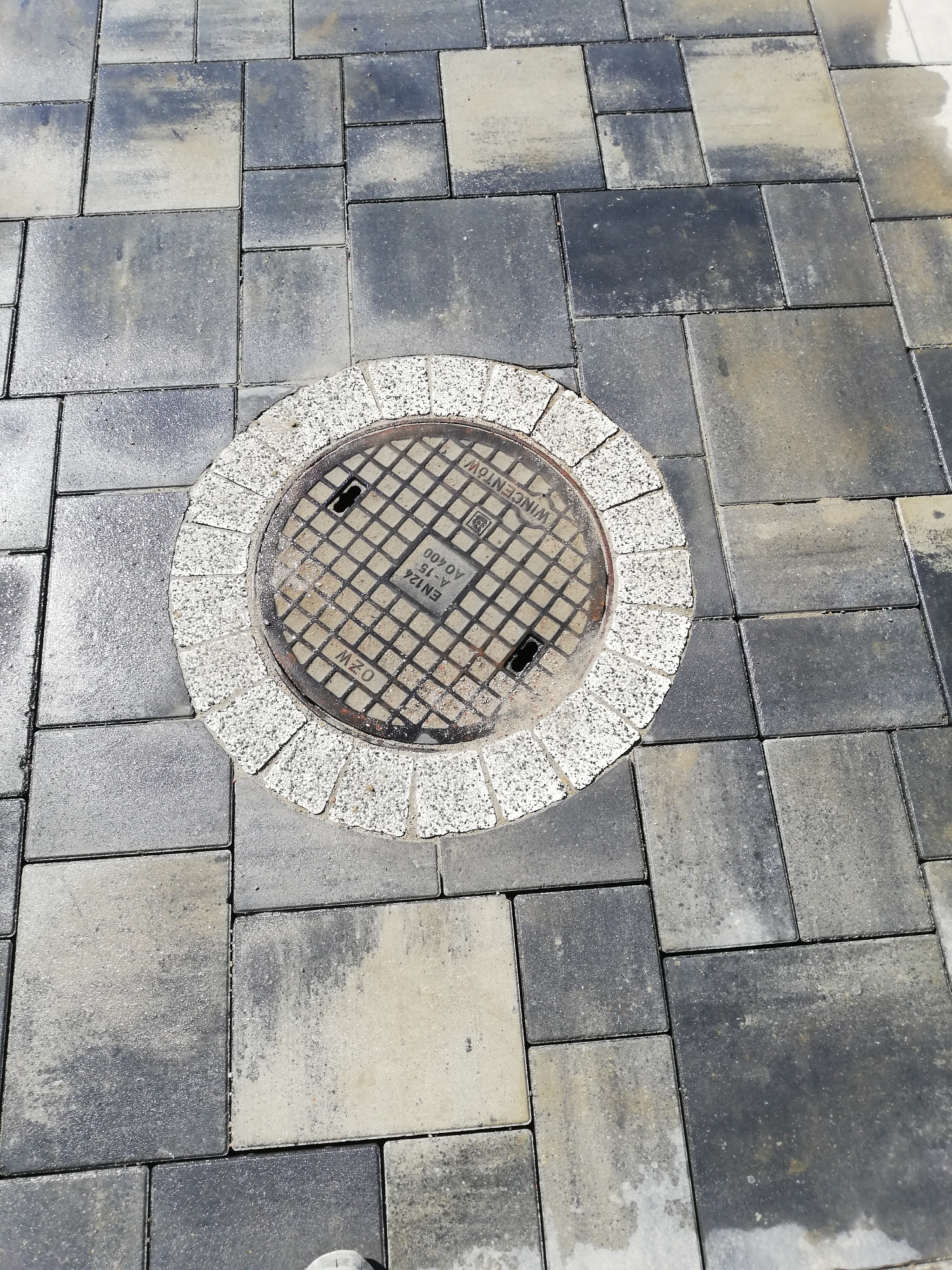 Szare, prostokątne kostki brukowe ułożone wokół okrągłej studzienki kanalizacyjnej z jasnym, granitowym obramowaniem.