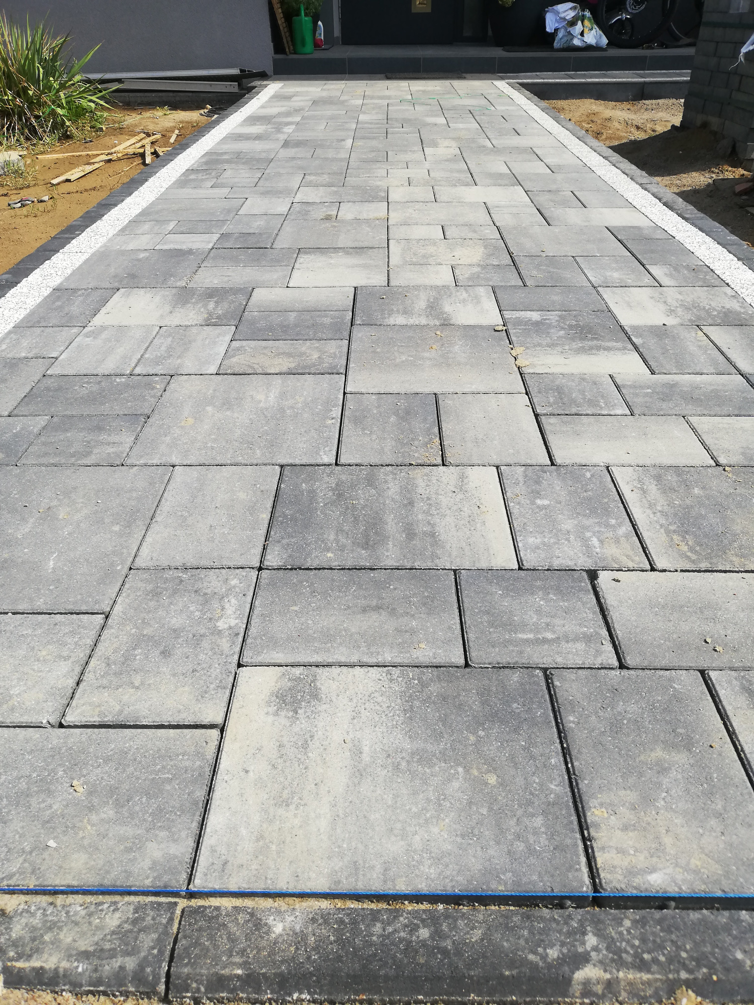 Ułożona szara kostka brukowa w różnych kształtach, obrzeża z jasnych kamyków, widok z perspektywy chodnika.