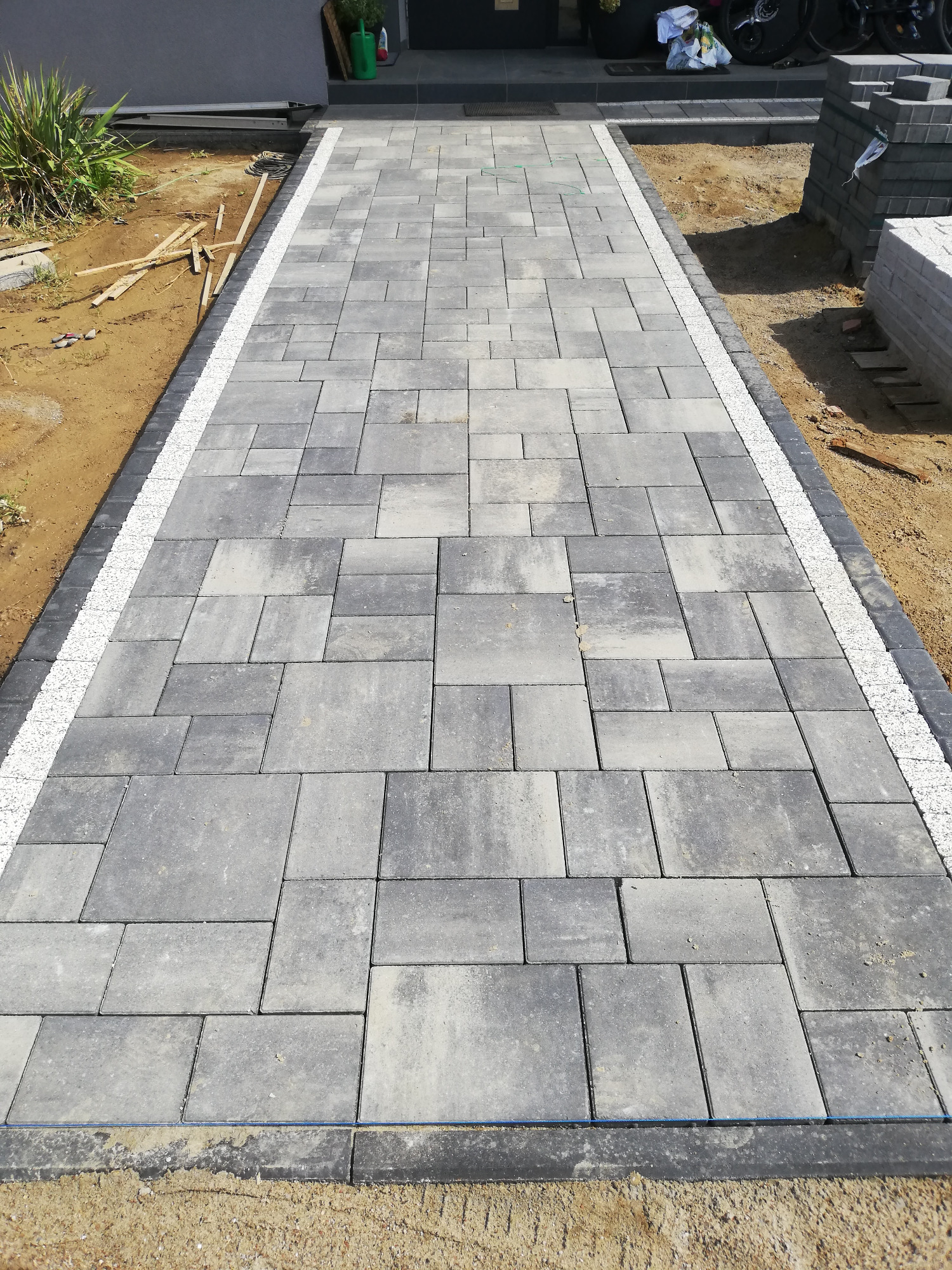 Świeżo ułożona, szara kostka brukowa prowadząca do domu, z ozdobnym pasem białego żwiru po bokach i widocznymi zapasami kostki po prawej stronie.