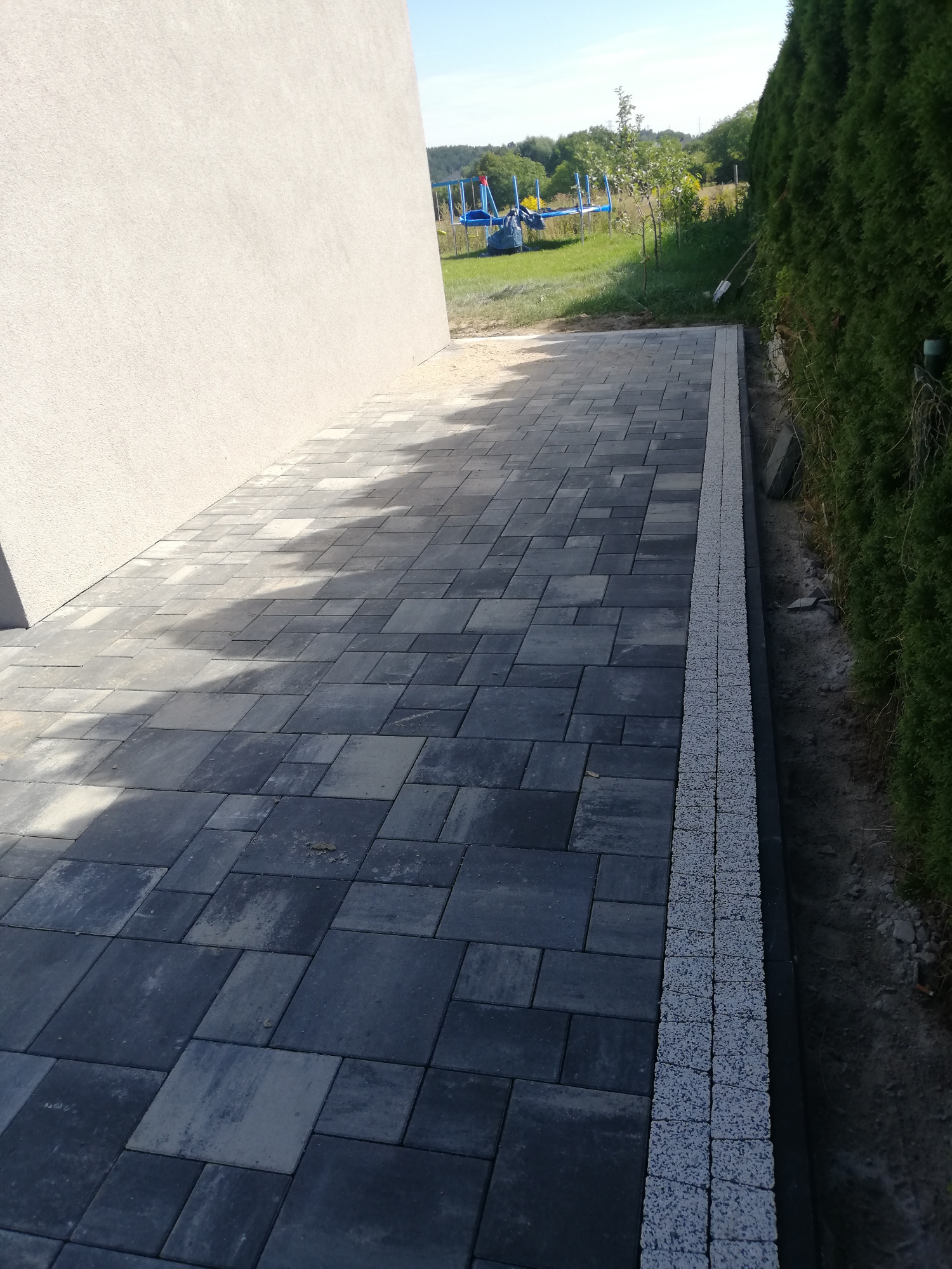 Ułożona, szara kostka brukowa z obrzeżem z jasnej kostki w Gorzowie Wielkopolskim, widok na fragment budynku i trawnik z trampoliną w tle.