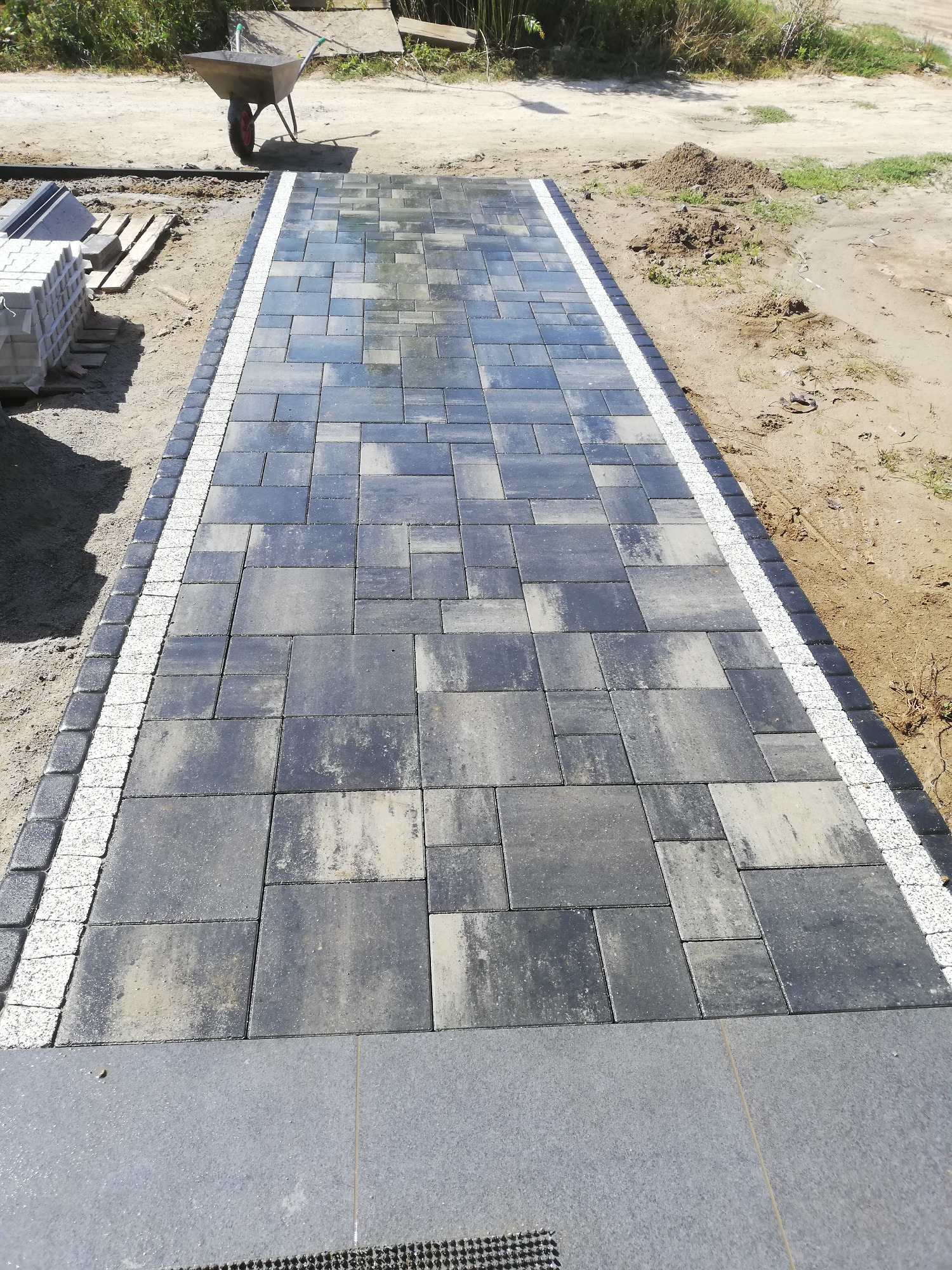 Świeżo ułożona, wilgotna kostka brukowa w odcieniach szarości, obramowana białym i ciemnoszarym obrzeżem, z taczką i stertą kostki w tle.