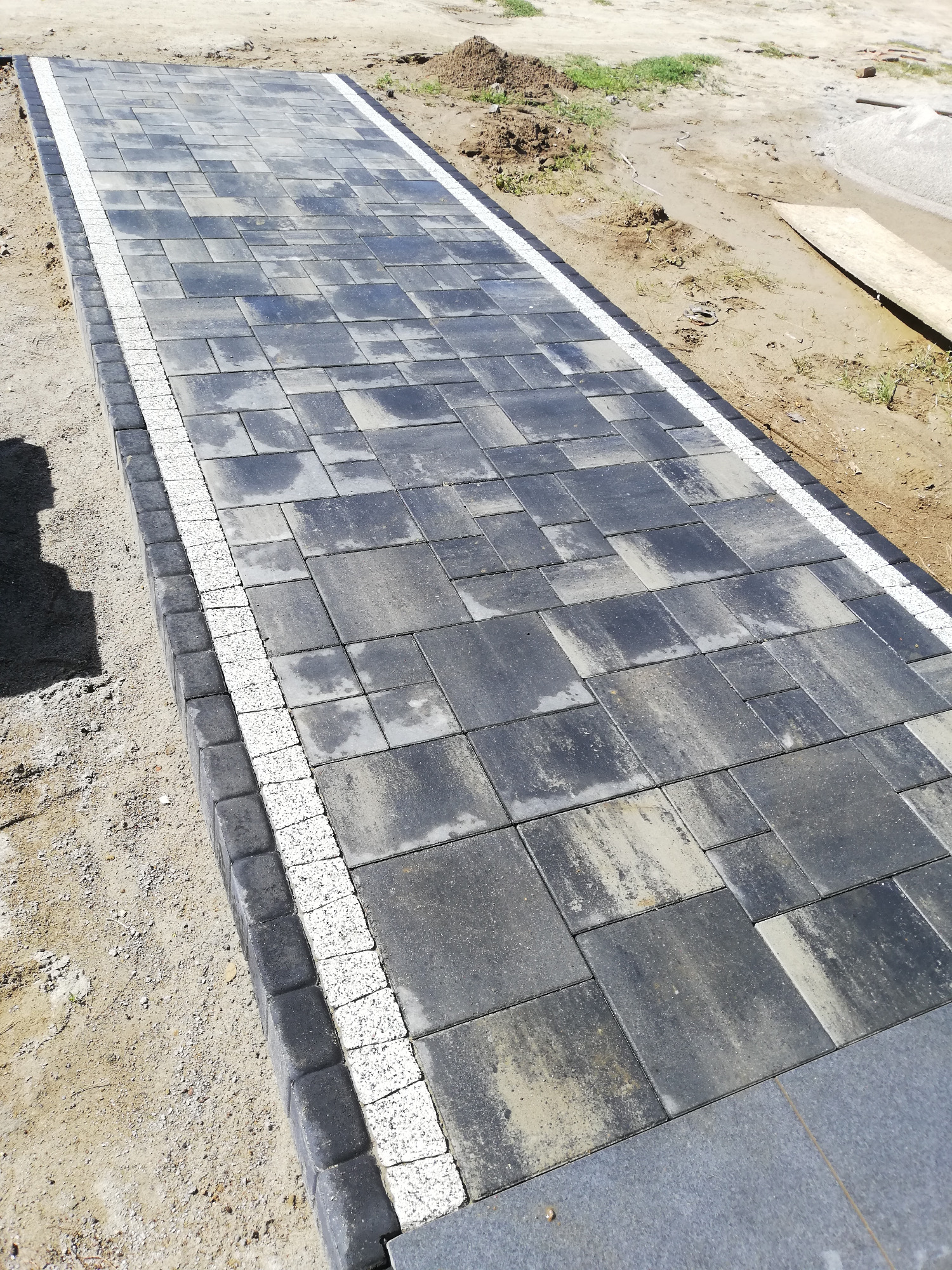 Ułożona ścieżka z kostki brukowej w odcieniach szarości, obrzeża z ciemnej kostki i białego grysu, widoczne ślady piasku po bokach.