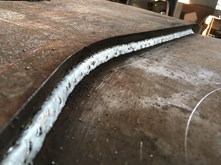 Świeżo wykonany spaw na zakrzywionej powierzchni metalowej, widoczne drobne odpryski spawalnicze i ślady po kredzie traserskiej.