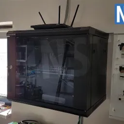 Szafa rack wisząca z osprzętem sieciowym i routerem na górze, obok skrzynka elektryczna, widok z boku, okno w tle.