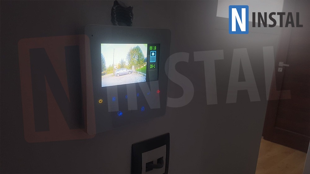 Zamontowany panel domofonu z widokiem na podjazd, na ekranie widoczny samochód z otwartym bagażnikiem, obok gniazdko sieciowe.