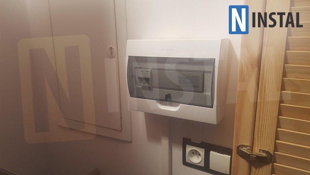 Zamknięta biała skrzynka bezpiecznikowa Schneider Electric na ścianie, obok gniazdka elektrycznego i drewnianych żaluzji.