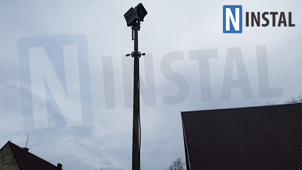 Słup oświetleniowy z dwoma reflektorami, zamontowany na zewnątrz, widoczny na tle pochmurnego nieba i fragmentów dachów budynków.