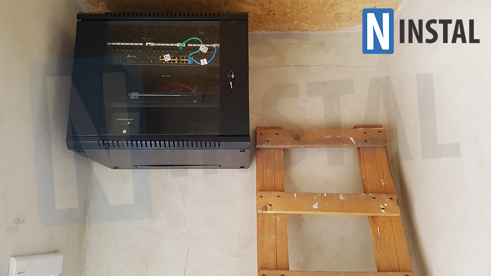 Zamknięta szafa rackowa z zainstalowanym switchem i rejestratorem na ścianie, obok drewniana drabina, surowy tynk.