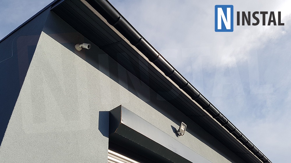 Zewnętrzna kamera monitoringu zamontowana na szarej elewacji budynku, widoczny system rynnowy i oświetlenie zewnętrzne, logo firmy w prawym górnym rogu.