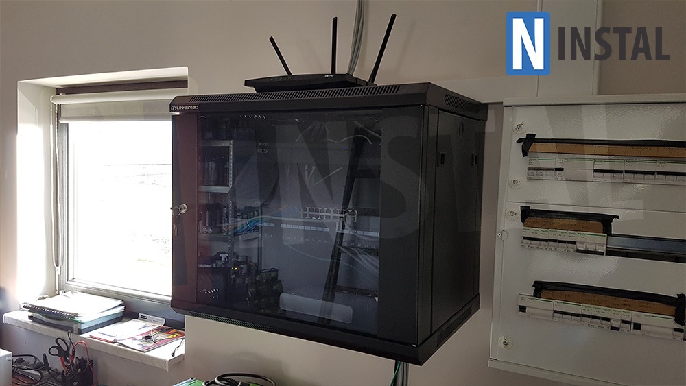 Szafa rack wisząca z osprzętem sieciowym i routerem na górze, obok skrzynka elektryczna, widok z boku, okno w tle.