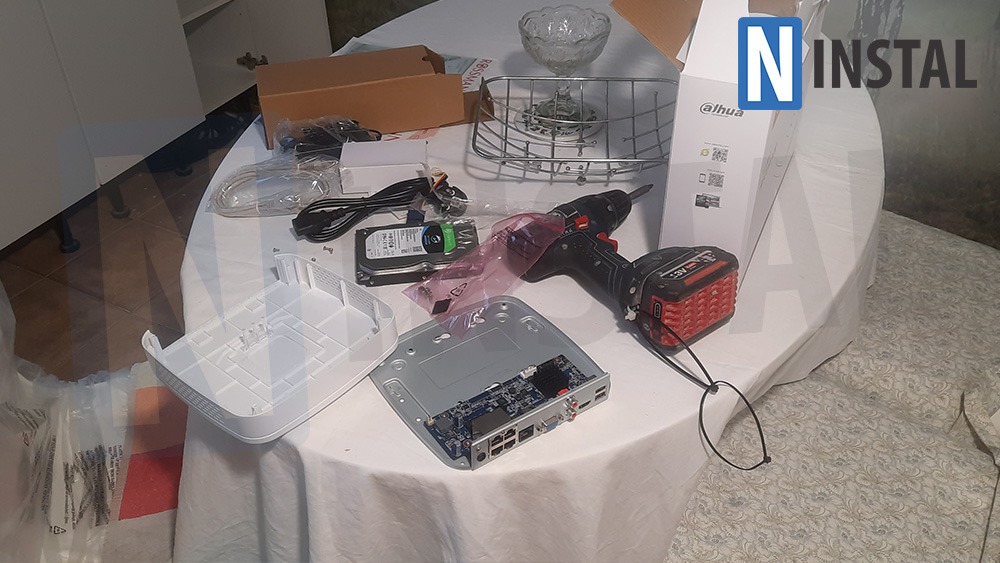 Rozpakowany rejestrator cyfrowy, dysk twardy, wkrętarka akumulatorowa i akcesoria na stole przykrytym białym obrusem podczas przygotowań do instalacji systemu monitoringu.