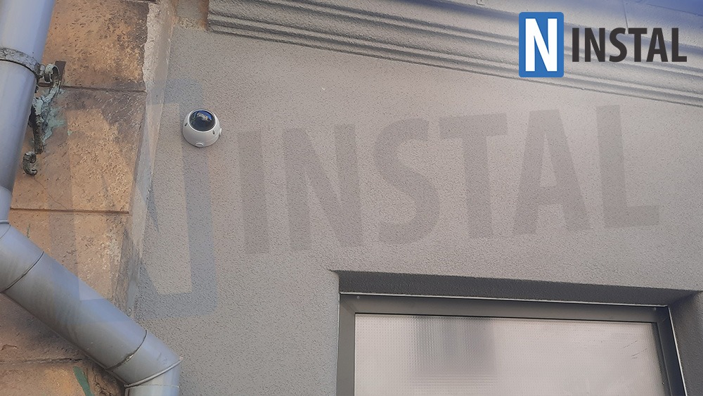 Kamera monitoringu kopułkowa zamontowana na szarej elewacji budynku obok rury spustowej, z widocznym logo firmy instalacyjnej w prawym górnym rogu.