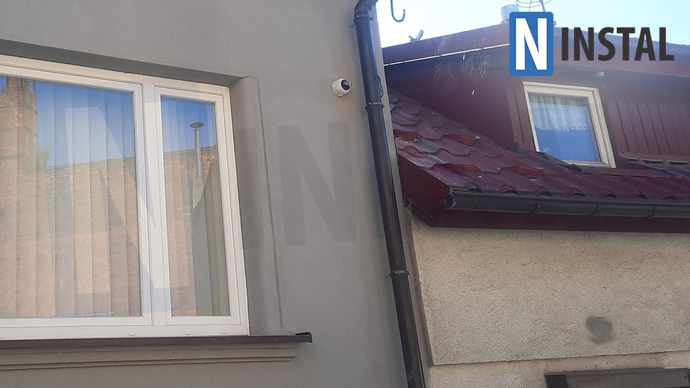 Kamera monitoringu zamontowana na szarej elewacji budynku obok okna, widoczny fragment dachu z rynną.