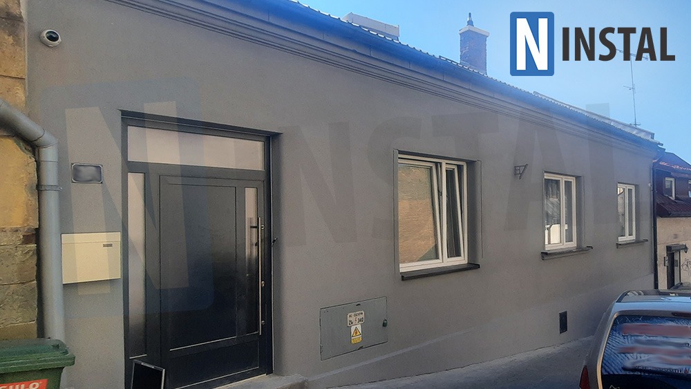 Szary budynek jednorodzinny z czarnymi oknami i drzwiami, widoczna kamera monitoringu nad drzwiami.