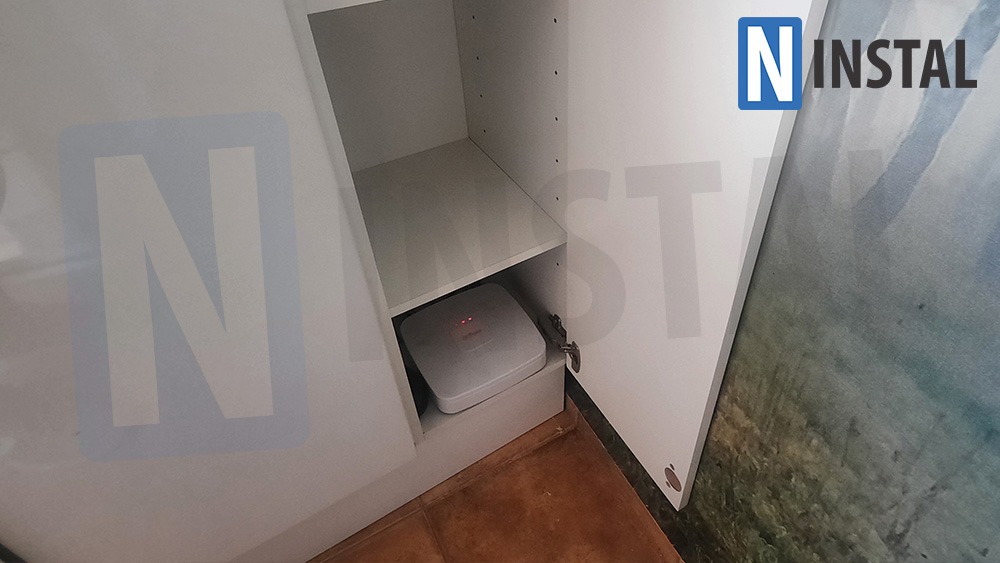 Biały punkt dostępowy Wi-Fi umieszczony w szafce z półkami; widoczne diody sygnalizacyjne i logo producenta; na drzwiach szafki logo firmy instalacyjnej.