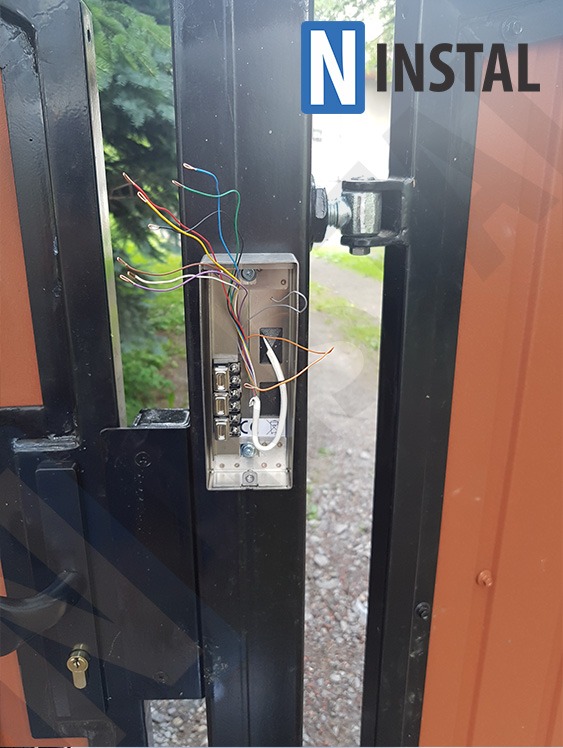 Interkom z rozpiętymi przewodami na czarnej metalowej bramie z brązowymi elementami, widoczne zielone drzewa w tle.