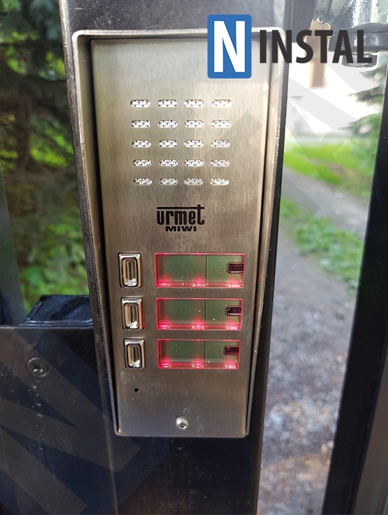 Zewnętrzny panel domofonowy Urmet Miwi ze stali nierdzewnej, zamontowany na czarnym metalowym słupku ogrodzenia, widoczne podświetlane przyciski i klawiatura numeryczna, w tle zieleń i chodnik.