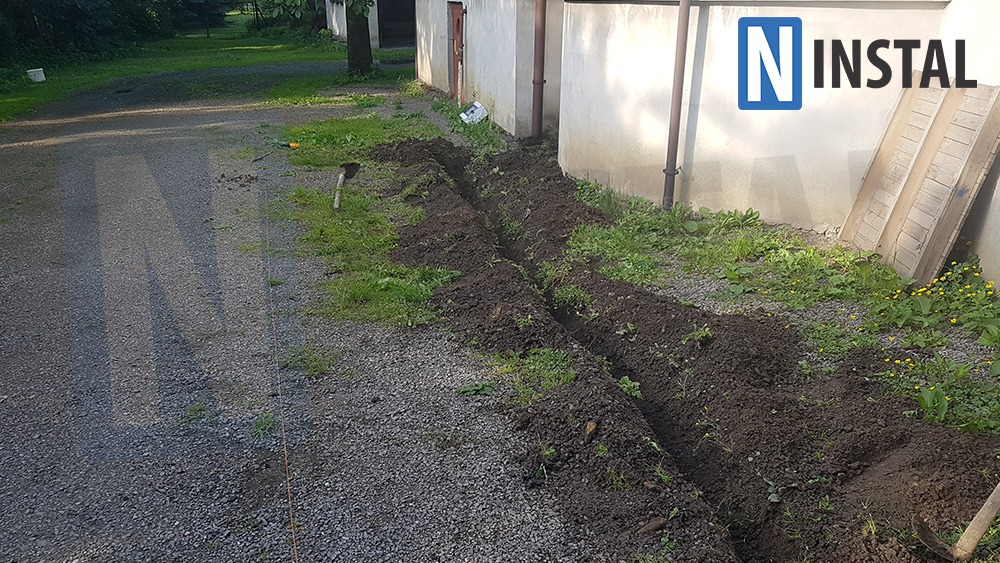Wykopany rów w ziemi, prawdopodobnie pod kable, widoczny fragment budynku i narzędzie w tle, logo firmy w prawym górnym rogu.