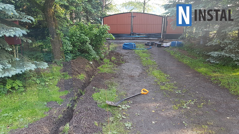 Wykopany rów w ziemi na posesji, widoczna łopata z pomarańczowym uchwytem, niebieski zwój kabla i brama w tle, logo firmy w prawym górnym rogu.