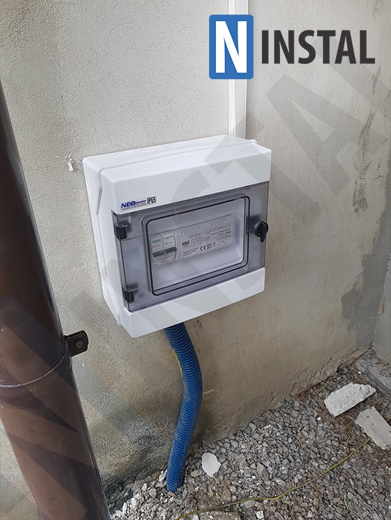 Zamontowana biała skrzynka elektryczna NEO IP65 z niebieską rurą karbowaną na szarej ścianie zewnętrznej, widoczne zabezpieczenia wewnątrz.