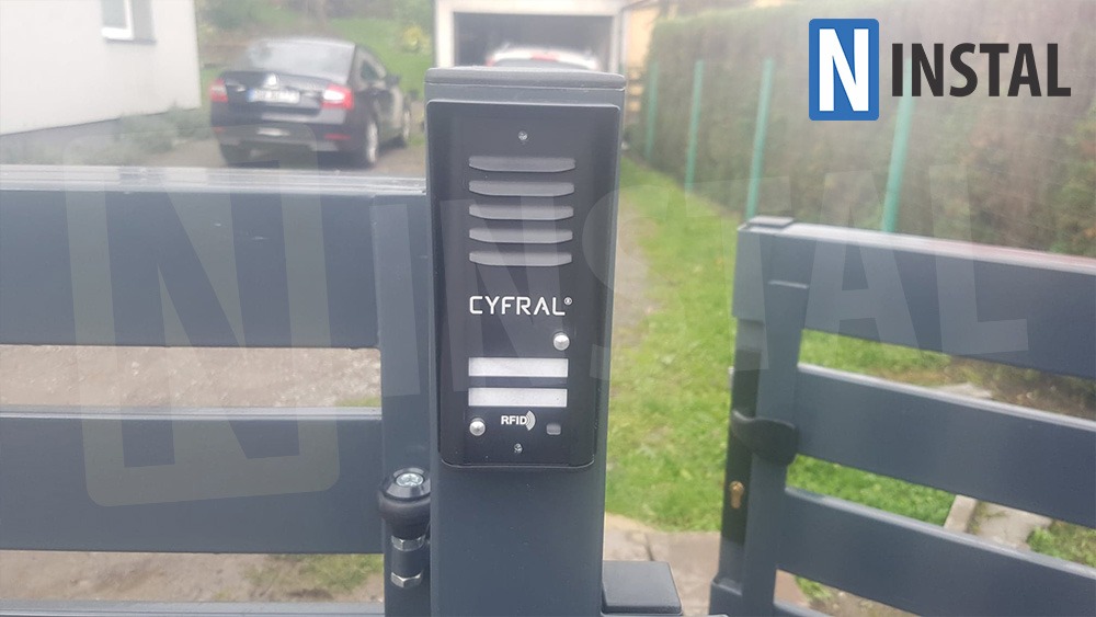 Czarny domofon Cyfral z czytnikiem RFID zamontowany na szarej metalowej bramie posesji z widocznym samochodem i garażem w tle.