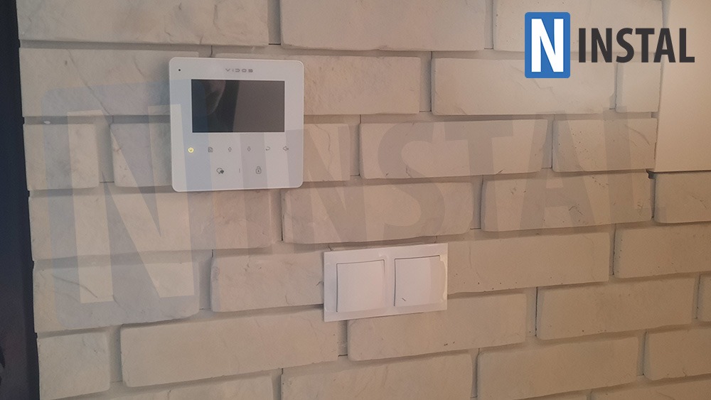 Biały panel dotykowy wideodomofonu Vidos zamontowany na dekoracyjnej ścianie z białej cegły, obok podwójnego włącznika światła.
