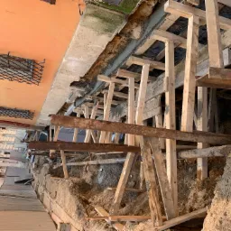 Wykop zabezpieczony drewnianą konstrukcją wsporczą i stalowymi belkami, widoczna odsłonięta ściana budynku z oknami zabezpieczonymi kratami, prace ziemne w trakcie realizacji.