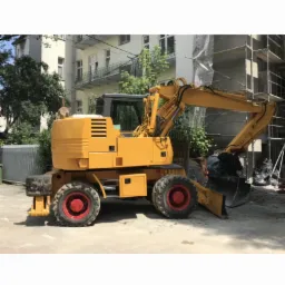 Żółta koparka kołowa z czerwonymi felgami stoi na utwardzonym placu budowy, w tle budynek w trakcie remontu z rusztowaniami.