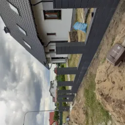 Częściowo ukończony, nowoczesny płot panelowy w kolorze grafitowym, widoczny z perspektywy gruntu, z domem w tle i stertą ziemi obok. Na ziemi leży brązowa obudowa automatyki do bramy.