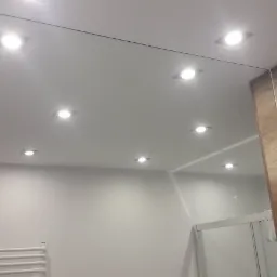 Nowoczesna łazienka z podwieszanym sufitem i wbudowanymi punktami świetlnymi, widoczne lustro i fragment szafki z drewnopodobnym wykończeniem, oraz grzejnik drabinkowy.