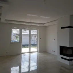 Wykończone wnętrze z beżowymi płytkami podłogowymi, białymi ścianami, nowoczesnym kominkiem z czarnymi akcentami, oknem balkonowym i dekoracyjnym sufitem z oświetleniem punktowym.