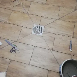 Układanie płytek podłogowych imitujących drewno, z centralnie umieszczonym odpływem liniowym, widoczne narzędzia: poziomica, nóż, pędzel i wiadro z zaprawą.